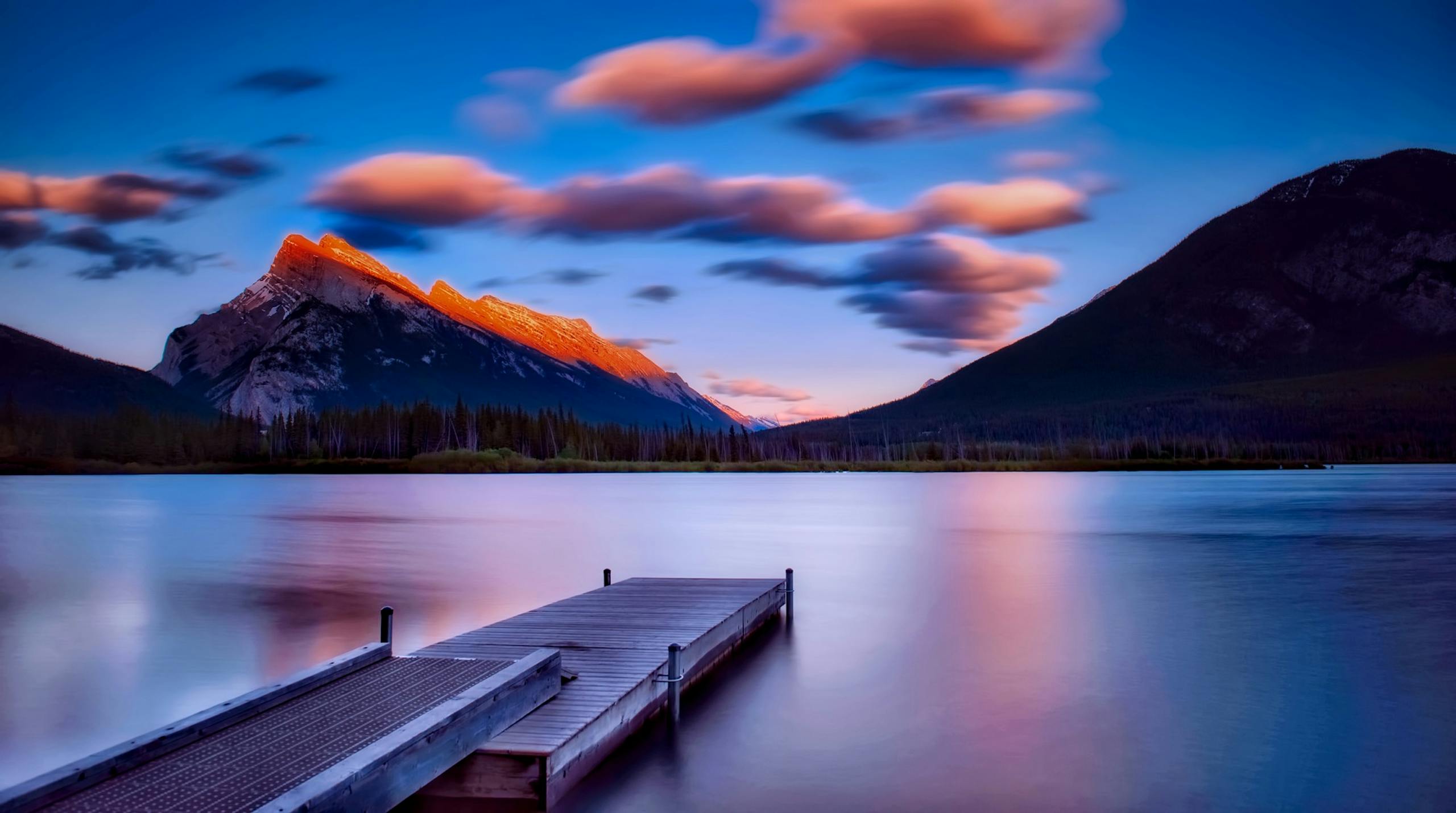 Breathtaking sunset view with mountains reflecting on a calm lake, featuring a wooden dock.