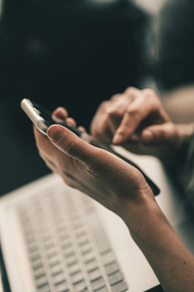 A person using a smartphone above a laptop keyboard in a workspace