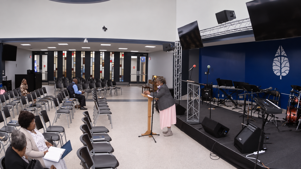 A church member speaks at a lectern beside seated congregants and the music area.