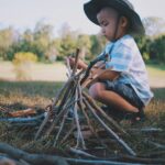 boy squat sitting beside wooden sticks formed to a pyramid during day