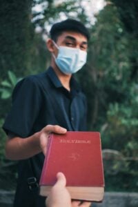 A young man wearing a face mask extends a red Holy Bible in an outdoor setting.
