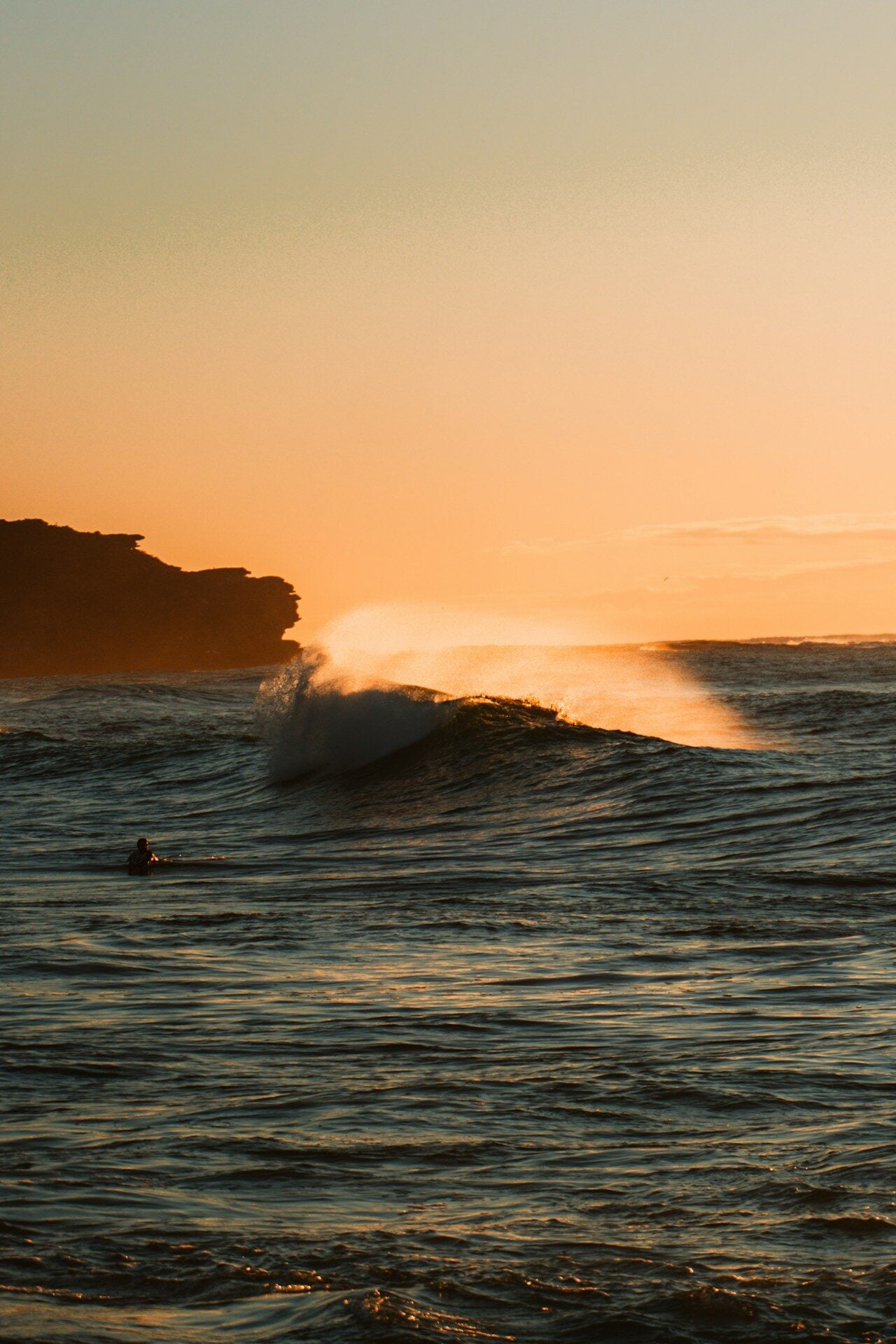 Surfer rides a wave at sunset near rocky coast.