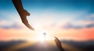 image of hand reaching up toward an extended hand reaching down. Sunlit cross is highlighted between the two hands.
