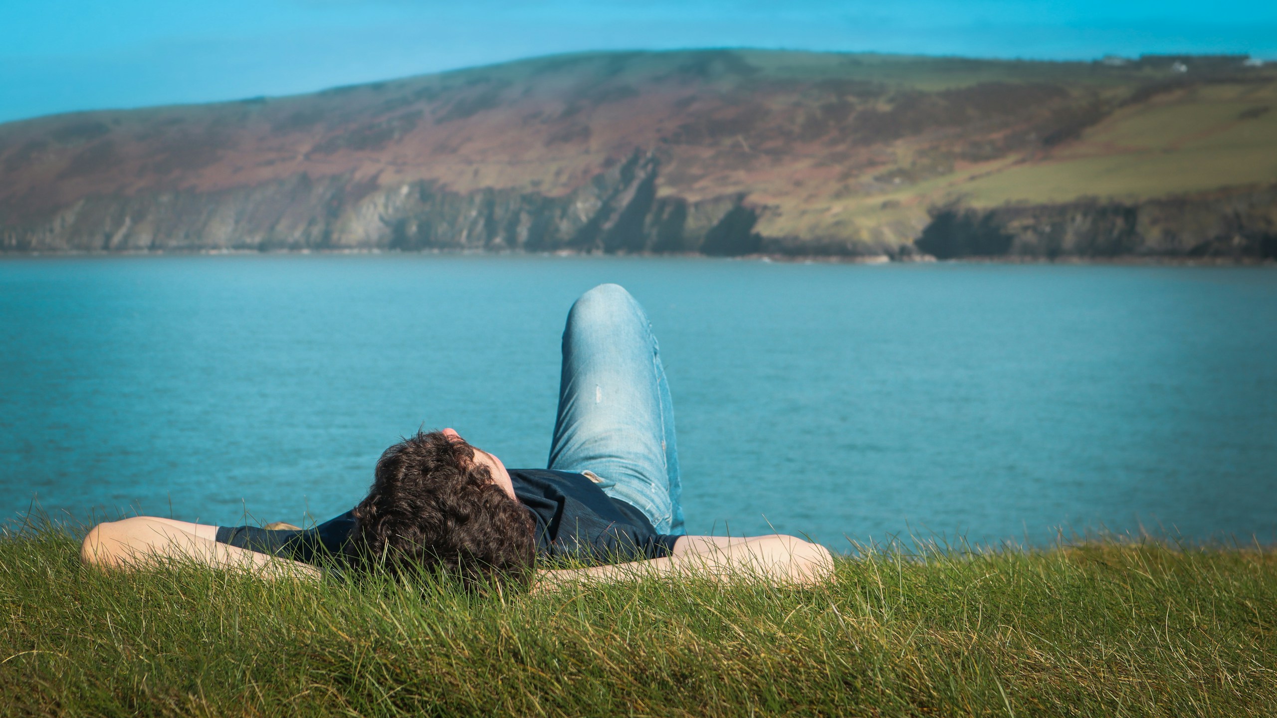 person in black shirt and blue denim jeans sitting on brown rock near body of water