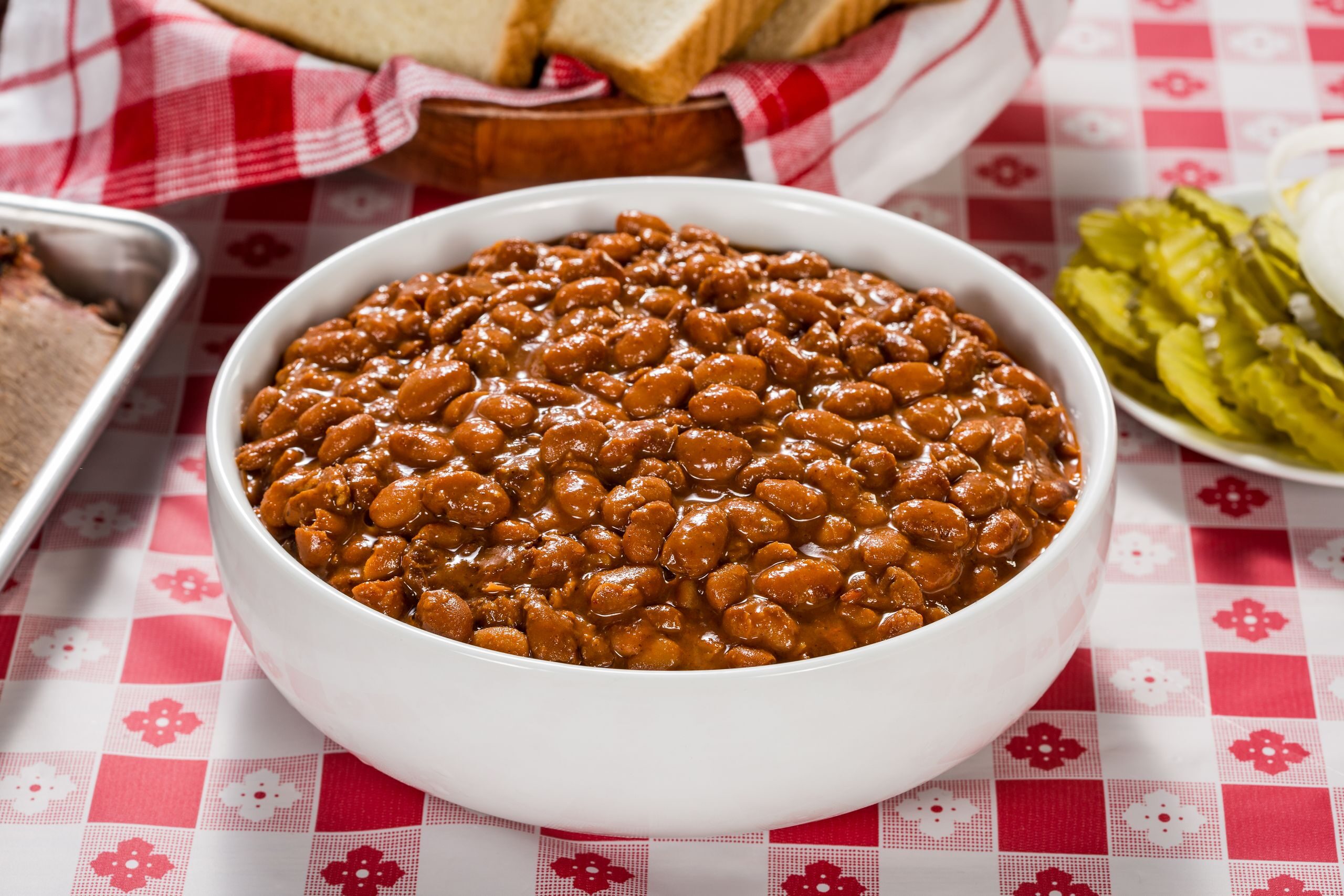 Close-up image of baked beans in a white bowl on a classic picnic table setting.
