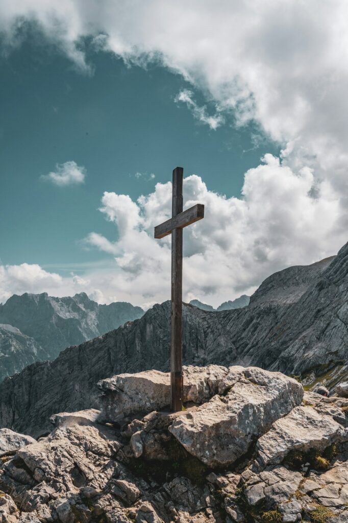 brown wooden cross on brown rock near green mountains under blue sky and white clouds during