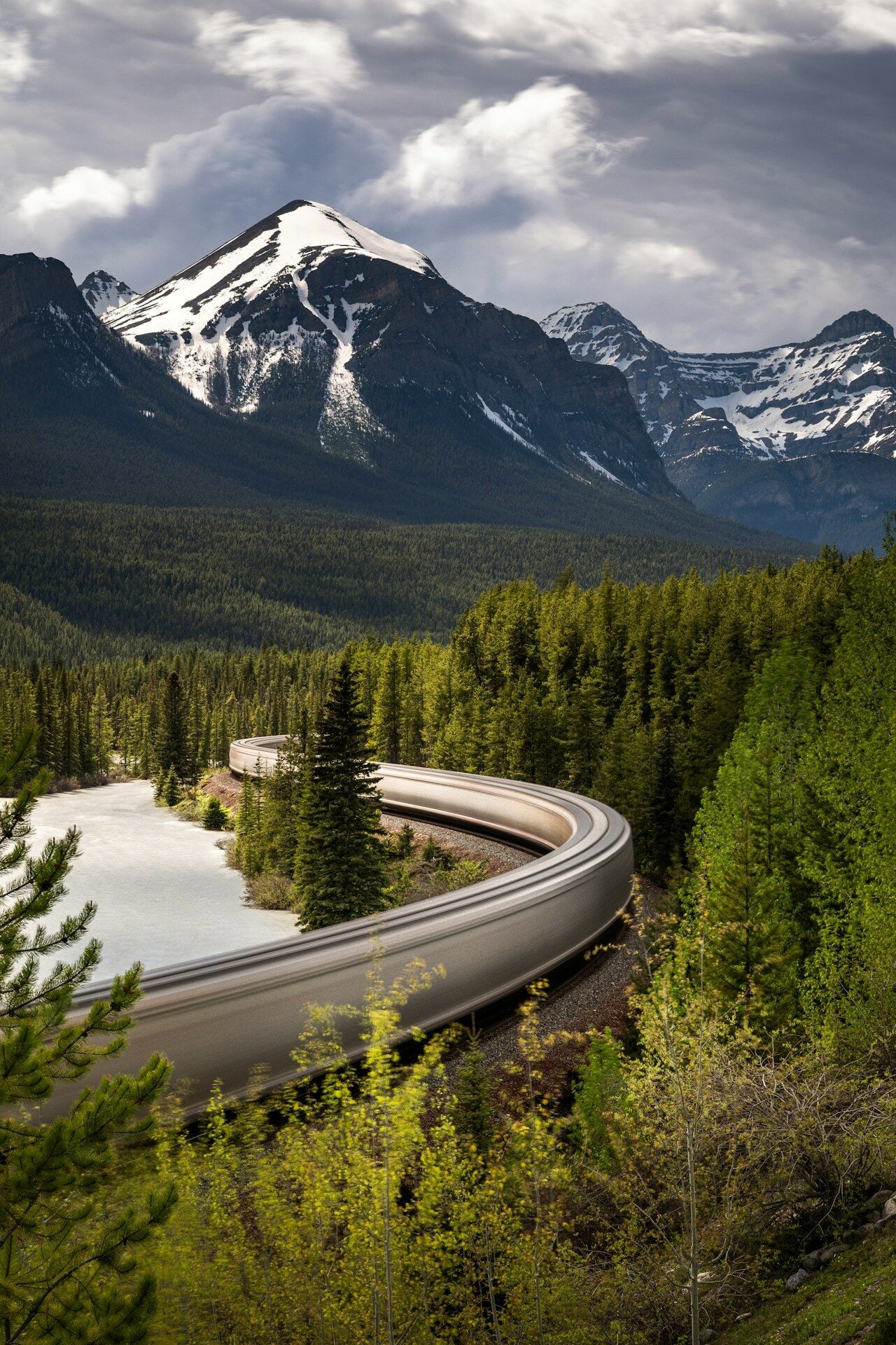 A train winds through a lush forest with snow-capped mountains.