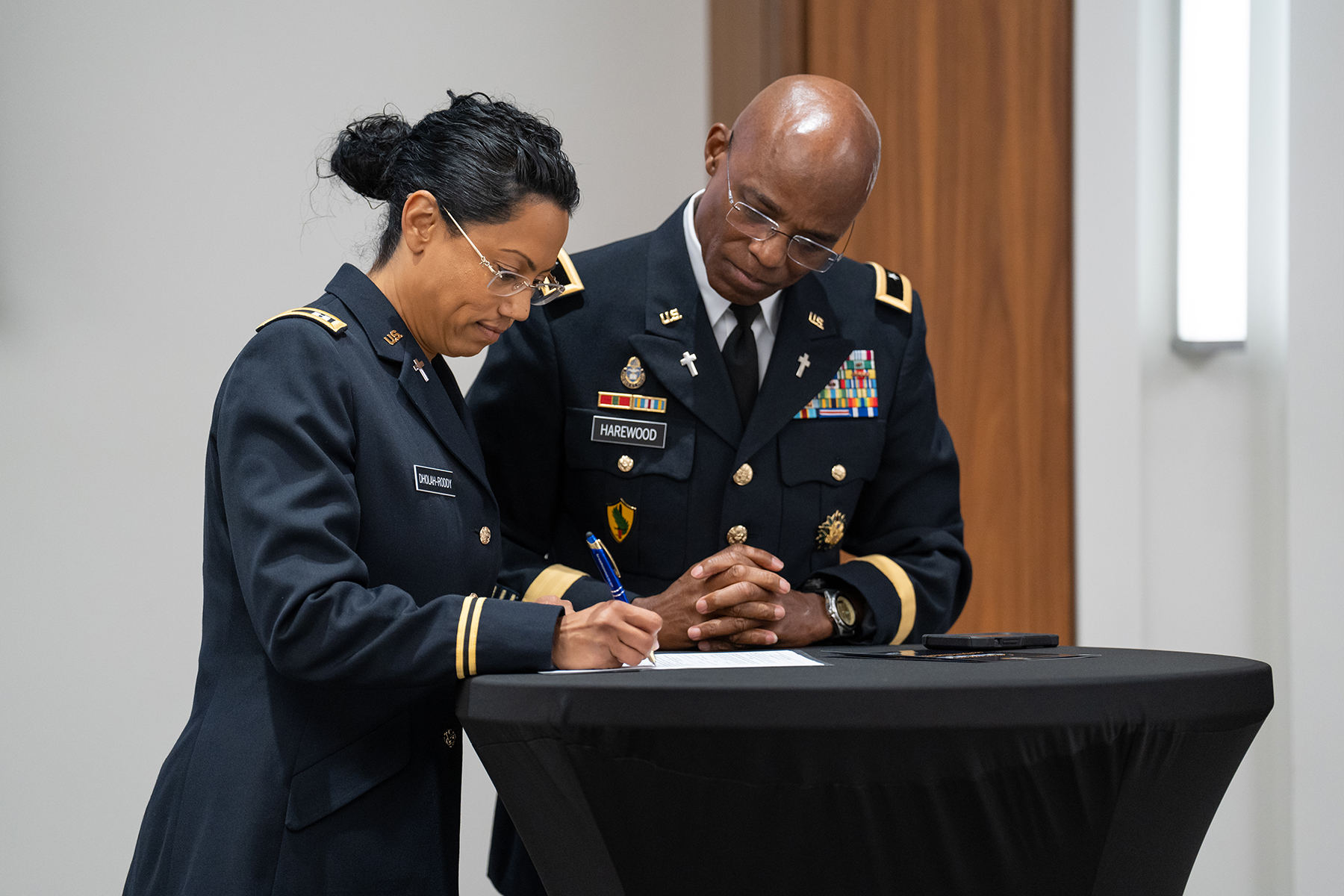 Woman with tanned skin signs a document as a black man watches. Both are in military uniform.