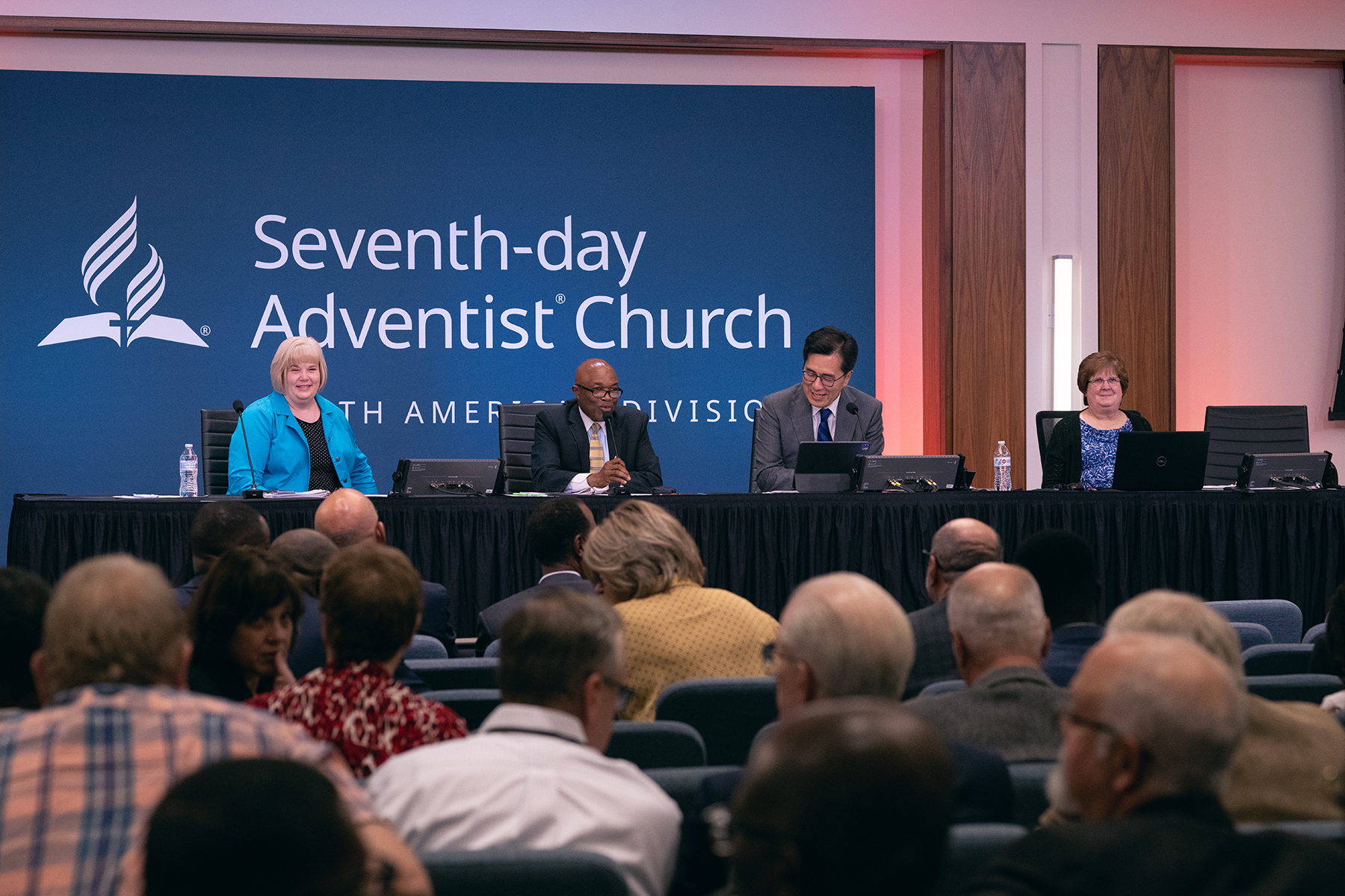 A table of four individuals seated at a table in front of an audience - L to R: white woman, Black man, Asian man, and white woman.