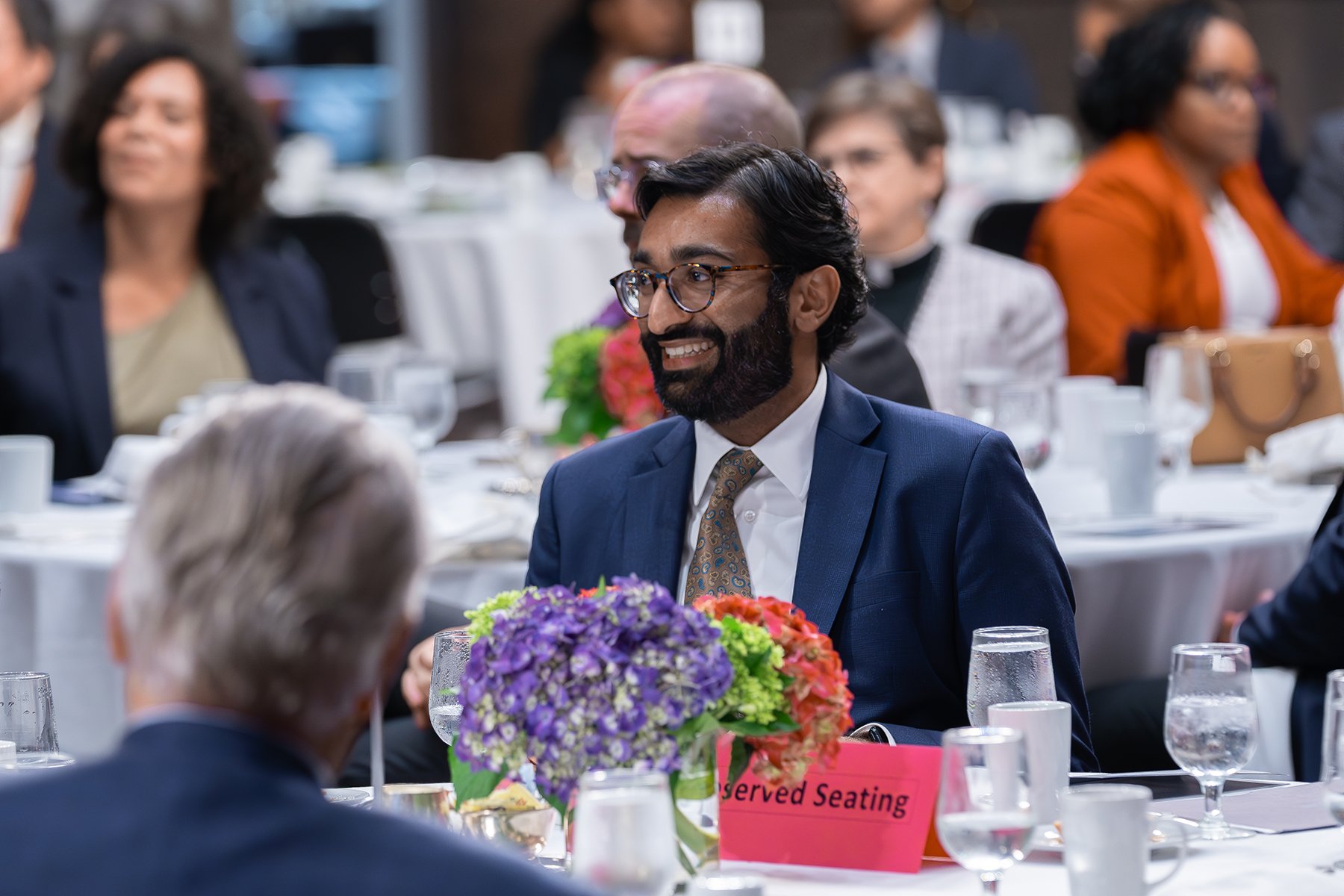 A smiling brown man is seated at a reserved seating table. People at other tables in background.