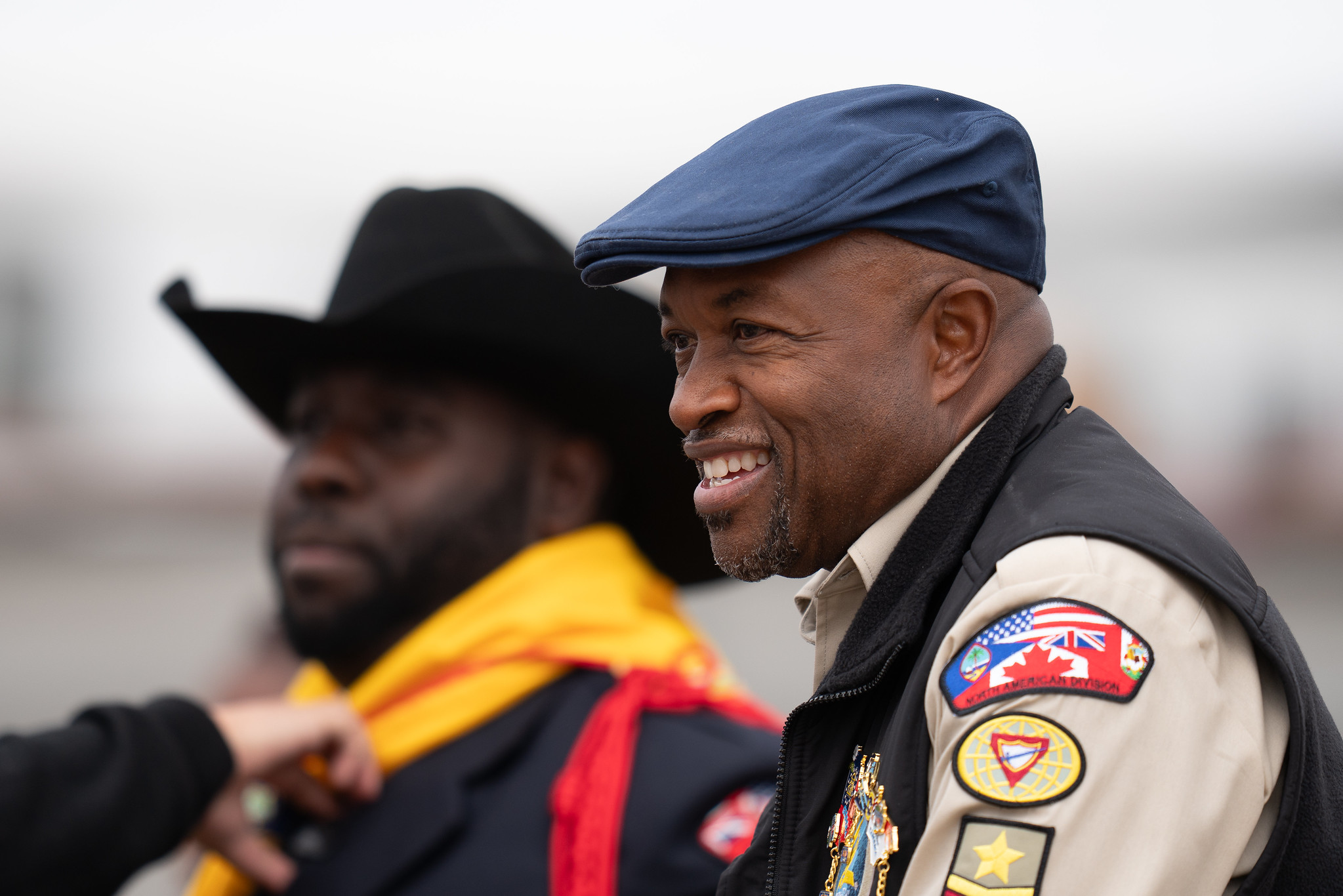 A closeup of two black men, one in focus, smiling, the other in the background with a serious expression. 