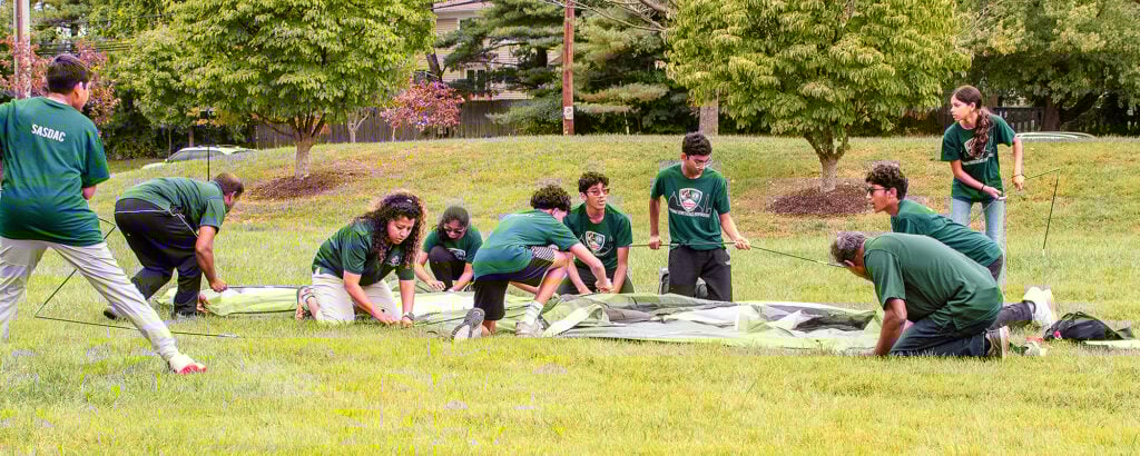 Young people pitch a tent on a grassy plain.