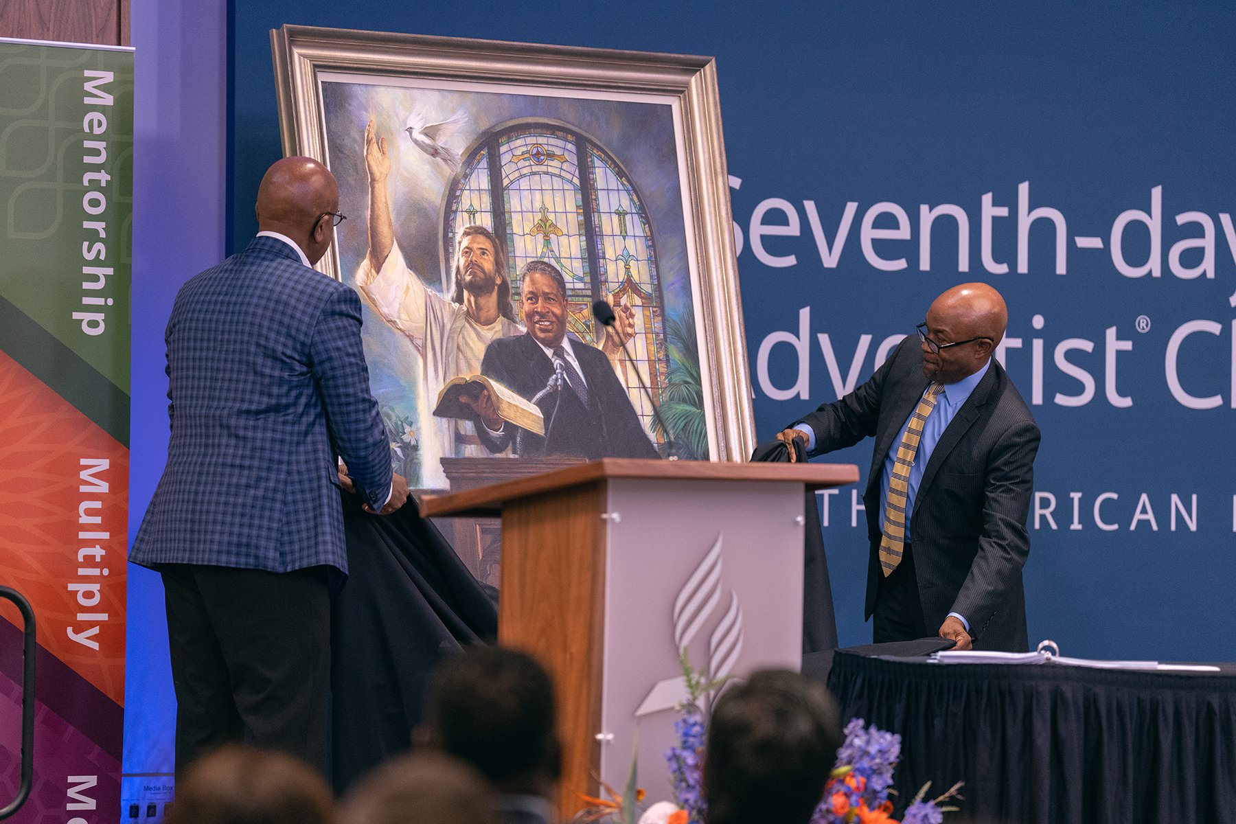 Two black men on a stage unveil a painting of a black man preaching.