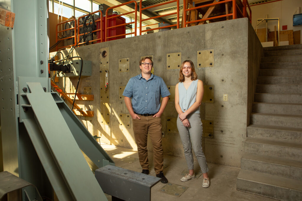 A young man and woman stand in the midst of an engineering lab, surrounded by building structures.