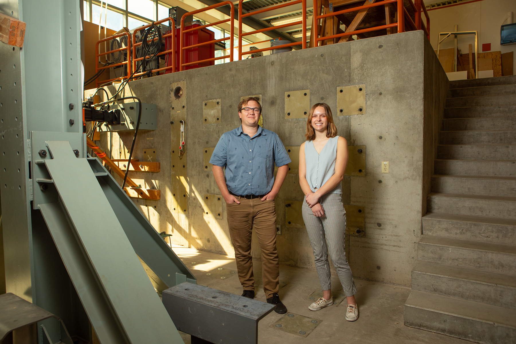 A young man and woman stand in the midst of an engineering lab, surrounded by building structures.