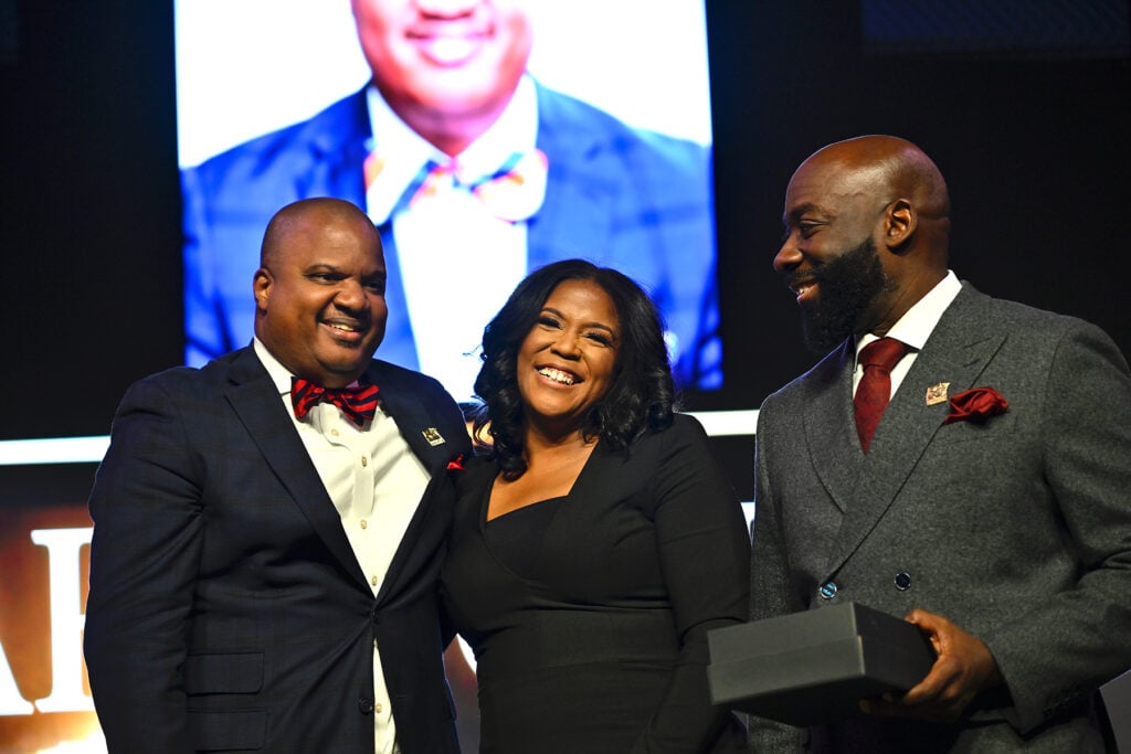a black man, black woman, and black man, smiling, on a stage in front of a big screen.