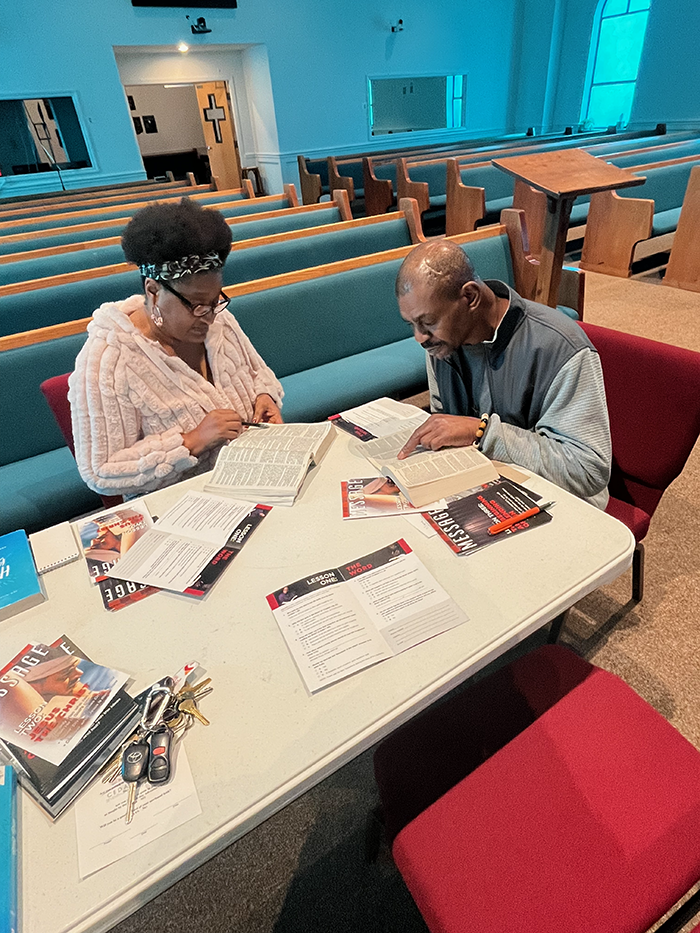 An African American woman studies the Bible at a table by a church pew with an African American man