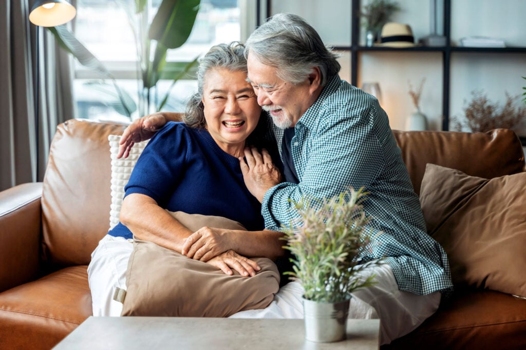 Older Asian couple shares a smile and hug while sitting on a couch.