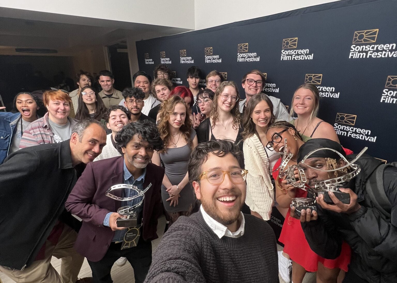 A diverse group of people stand in front of a media wall, some holding awards that look like a film strip