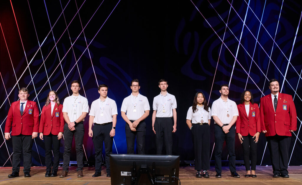 A diverse line-up of young men and women on a stage wearing white shirts, black pants and some with red jackets.