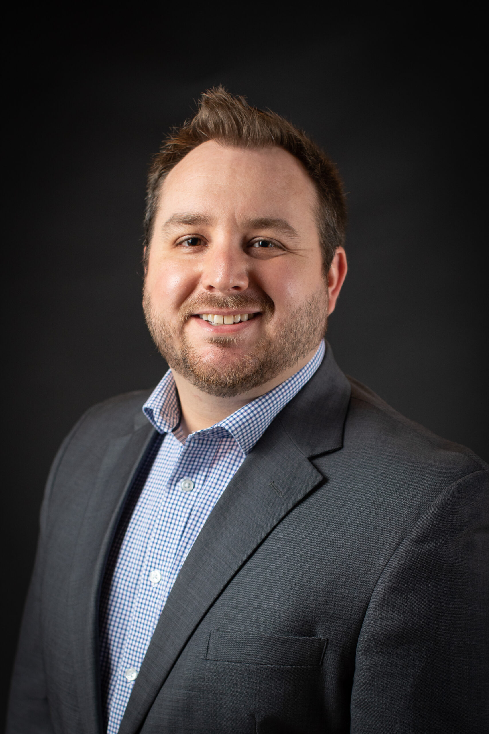 Headshot of a white man in a grey suit, with a blue and white checkered dress shirt