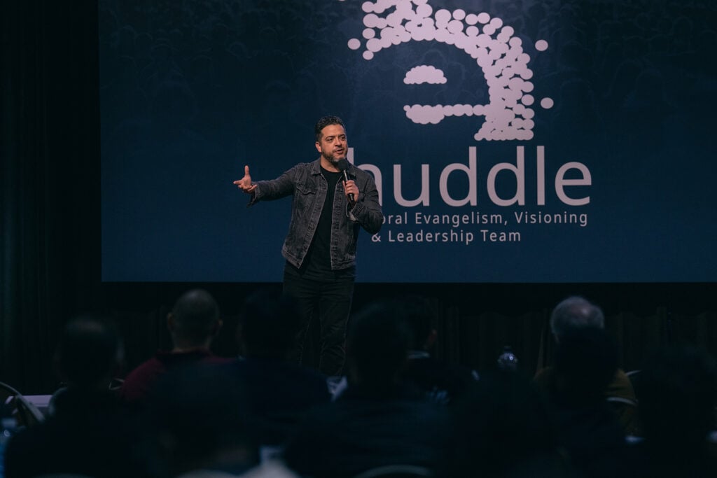 Hispanic man speaking on a stage with a screen reading eHuddle behind him.