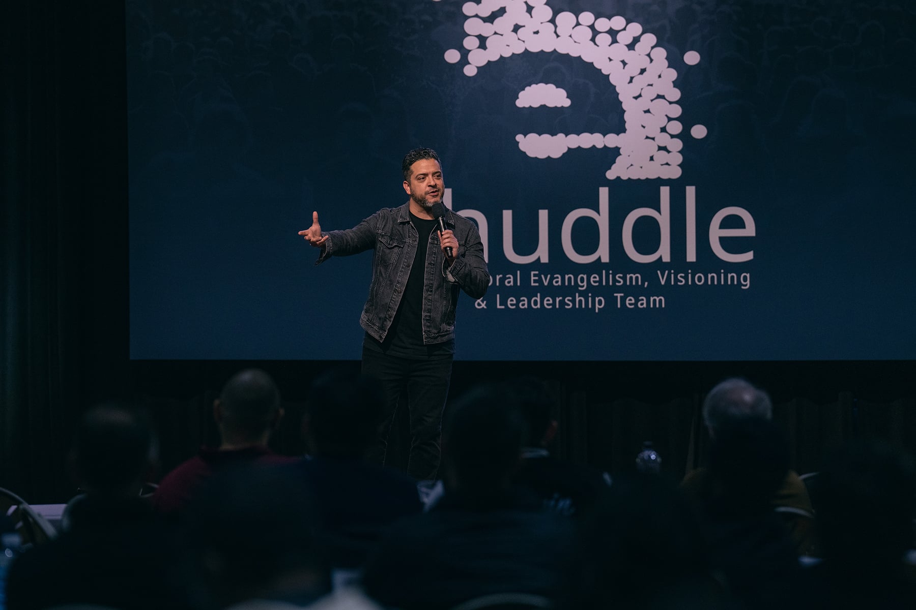 Hispanic man speaking on a stage with a screen reading eHuddle behind him.