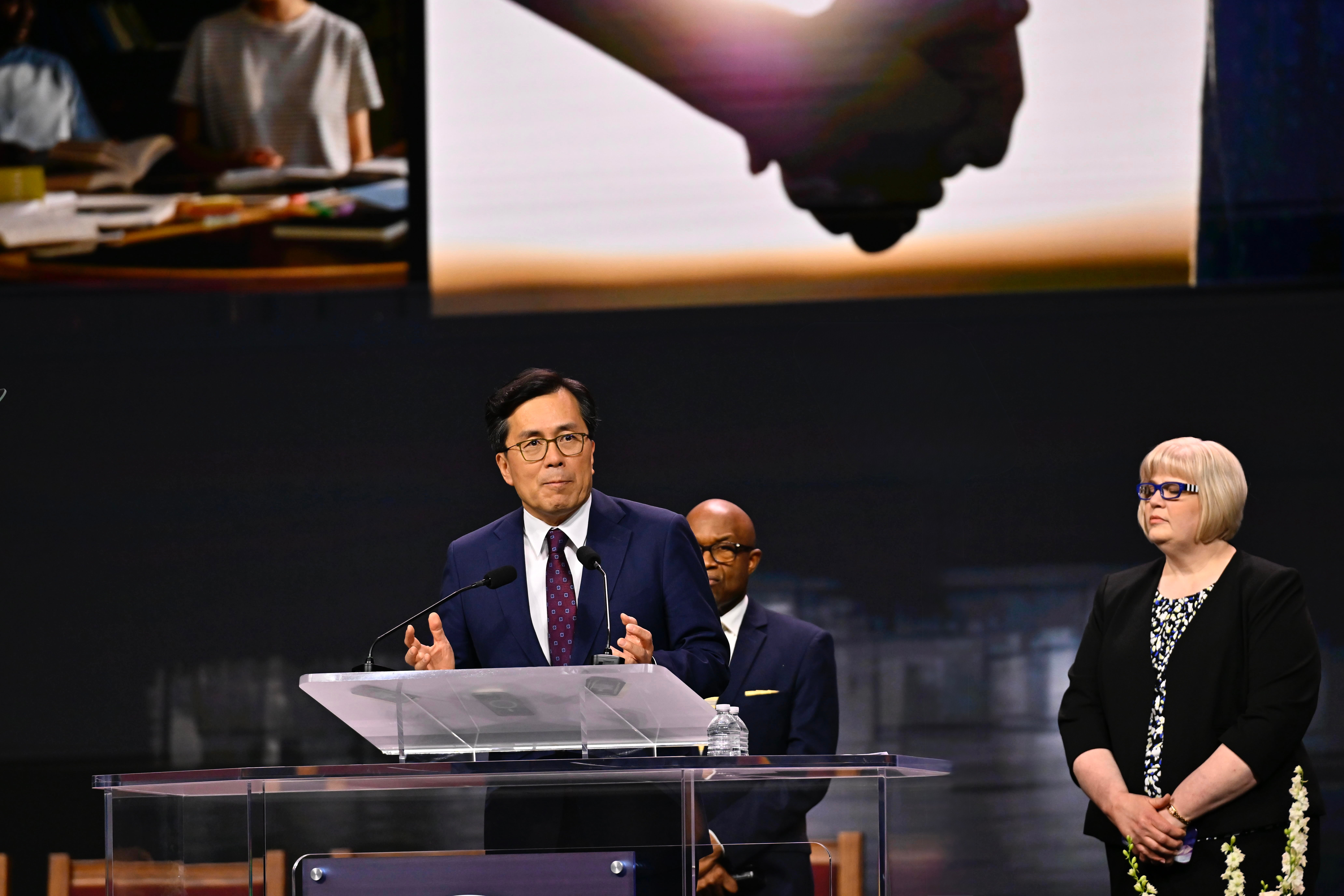 An Asian man speaks at a podium, with a black man and white woman in the background.