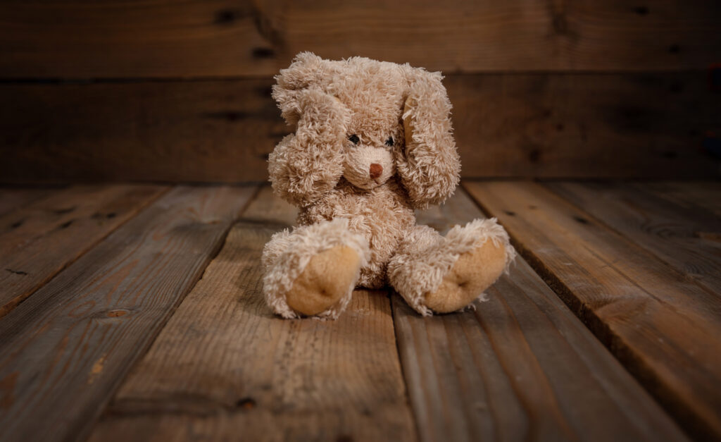 Teddy bear covering eyes while alone in an empty corner.