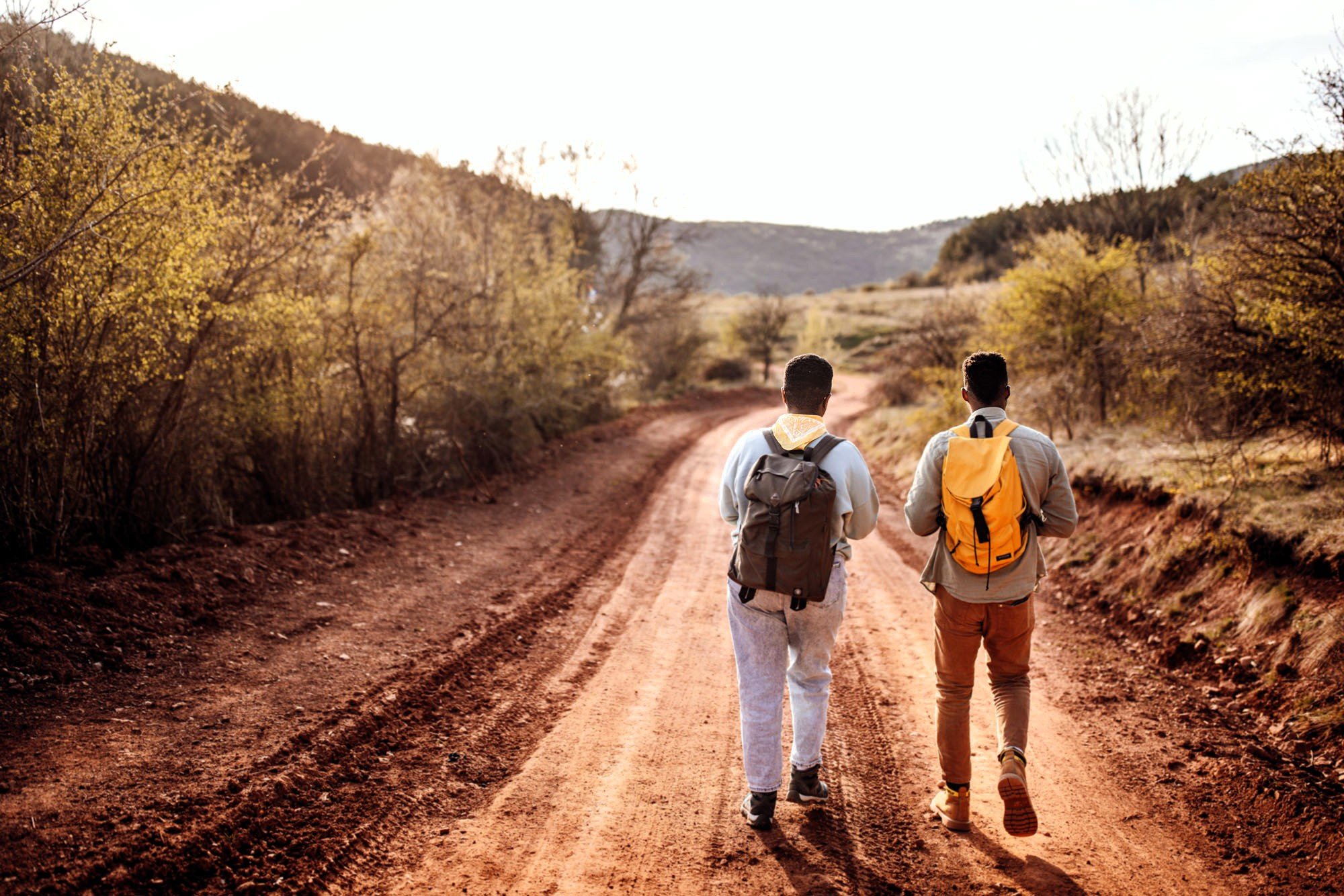 Two young men of color walking down a rural dirt road wearing daypacks