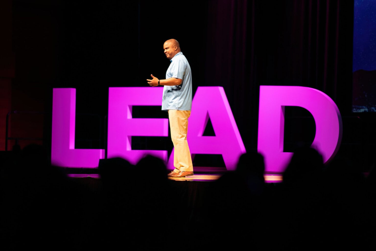 Conference speaker stands on stage in front of a large purple prop spelling out the word LEAD.