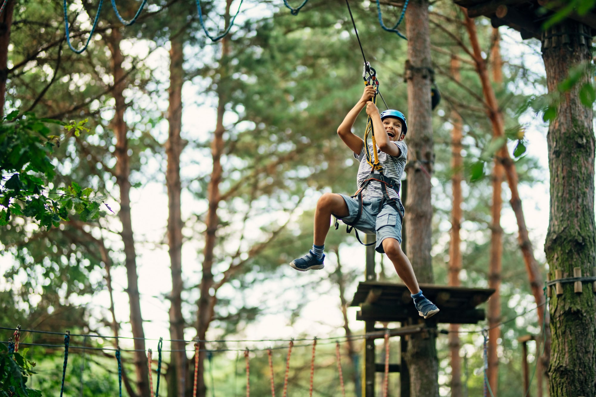 Young boy in a harness on a ropes course in the forest sliding along a cable to the next tree platform.