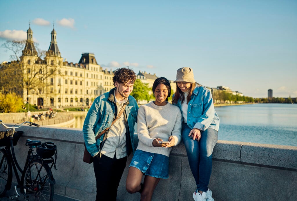 Three college age friends in Europe sit on the edge of a bridge in front of a ornate older building along a river.