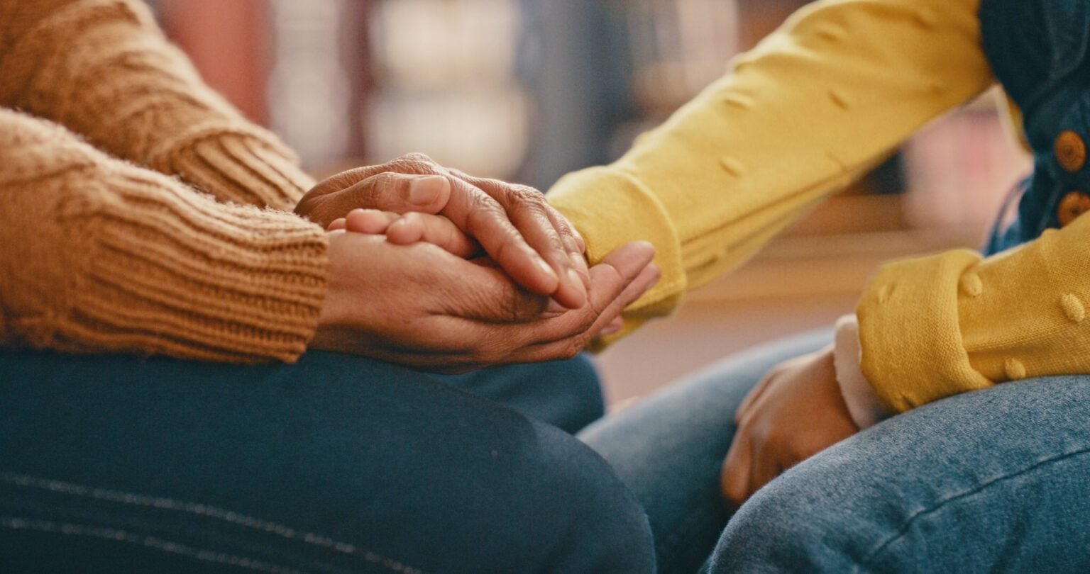 Close up photo of two people sitting knee to knee with one clasping the hand of the other to comfort them.