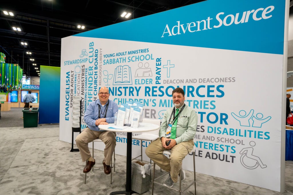 Two men sit on stools by their business display at a convention exhibit hall.