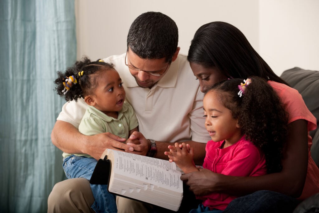 Young family with two children praying together at home on their couch. Toddler has eyes open and is looking at her mother.