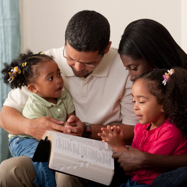 Young family with two children praying together at home on their couch. Toddler has eyes open and is looking at her mother.