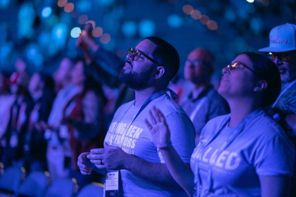 Photo focuses on two people in a large crowd at an event. People are in blue light from the stage and have their eyes closed and hands raised.