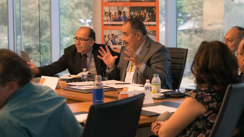 Group of people sitting around a conference table discussing ministry work. Sign in Spanish in the background.