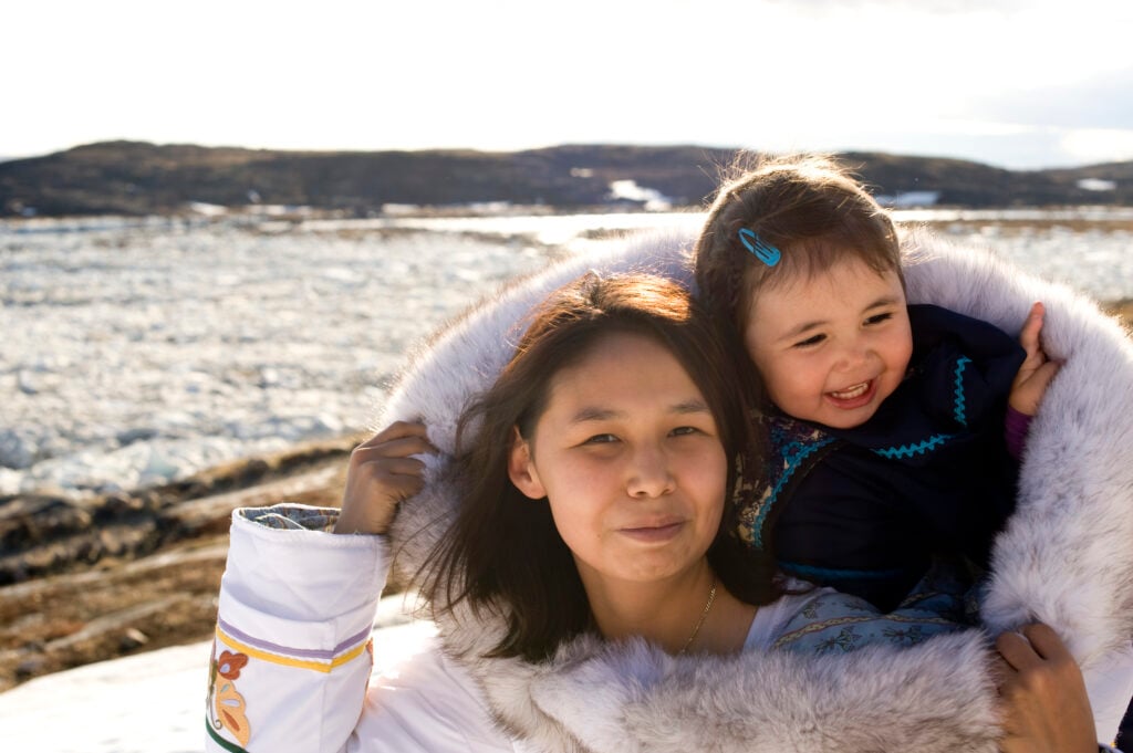 An Inuit mother has her daughter in her amouti, a traditional way to carry the young people. This is on the tundra of Baffin Island, Nunavut, Canada in late spring.
