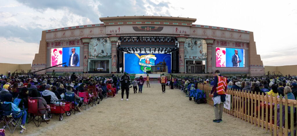 Photo showing a large outdoor stage for a religious yout program at dusk and many people sitting on camping chairs making up the audience.