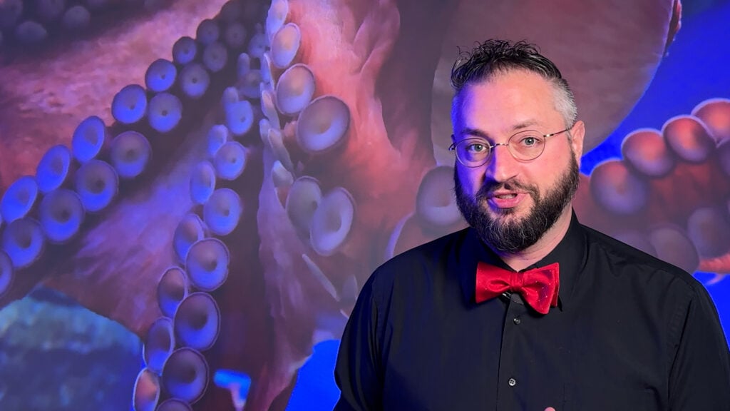 Caucasian man in black dress shirt and red bowtie poses in front of octopus picture