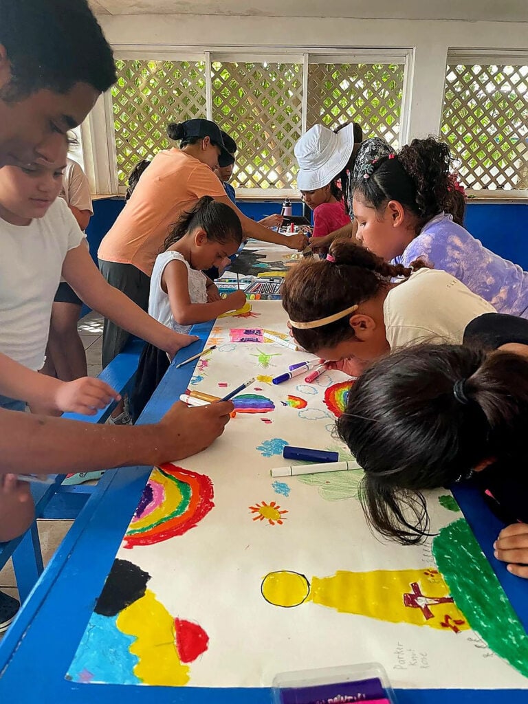 Children are pictured drawing on a long white paper with the help of volunteers. 