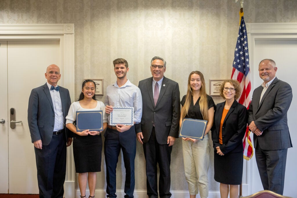 Three Southern Adventist University students stand alongside faculty with certificates, posed in front of the U.S. flag.