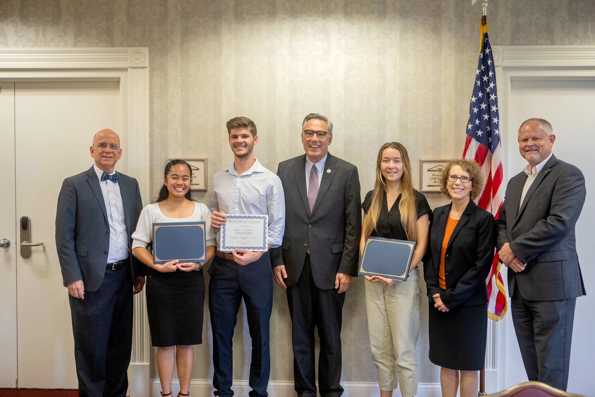 Three Southern Adventist University students stand alongside faculty with certificates, posed in front of the U.S. flag.