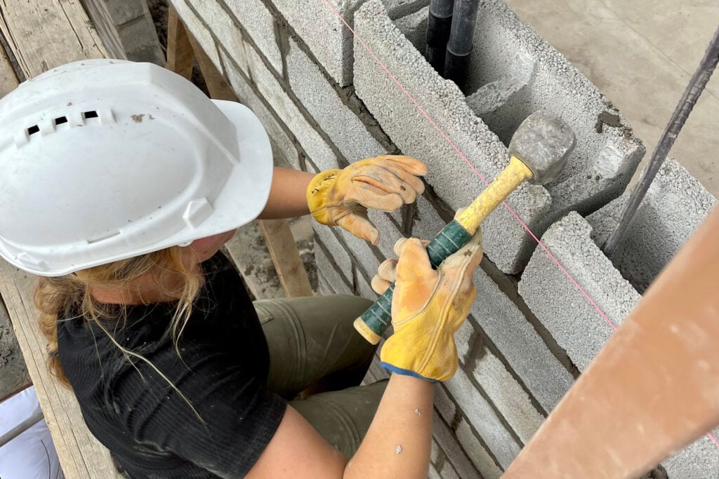 A young woman wearing a hard hat works on a concrete wall. 
