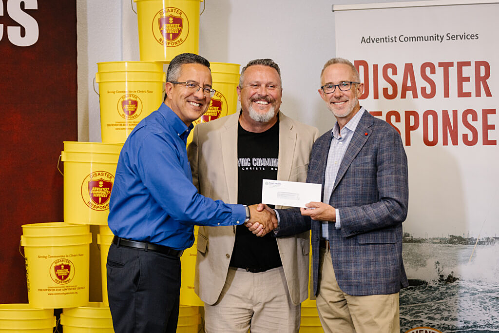 Three men smile as they present a donation check in front of yellow Adventist Community Services disaster response buckets.
