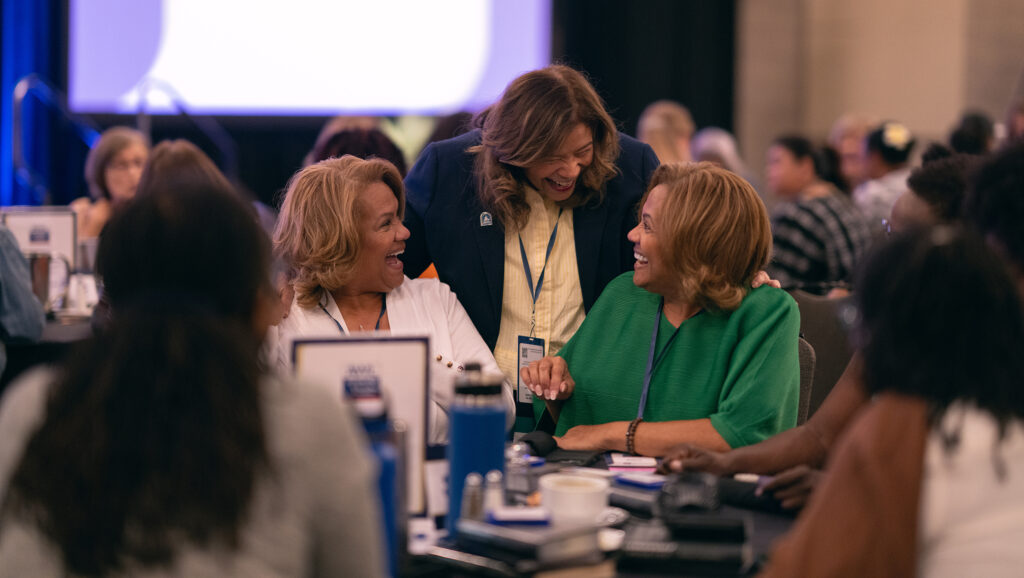 Three women, two seated at a table, one standing, share a laugh.