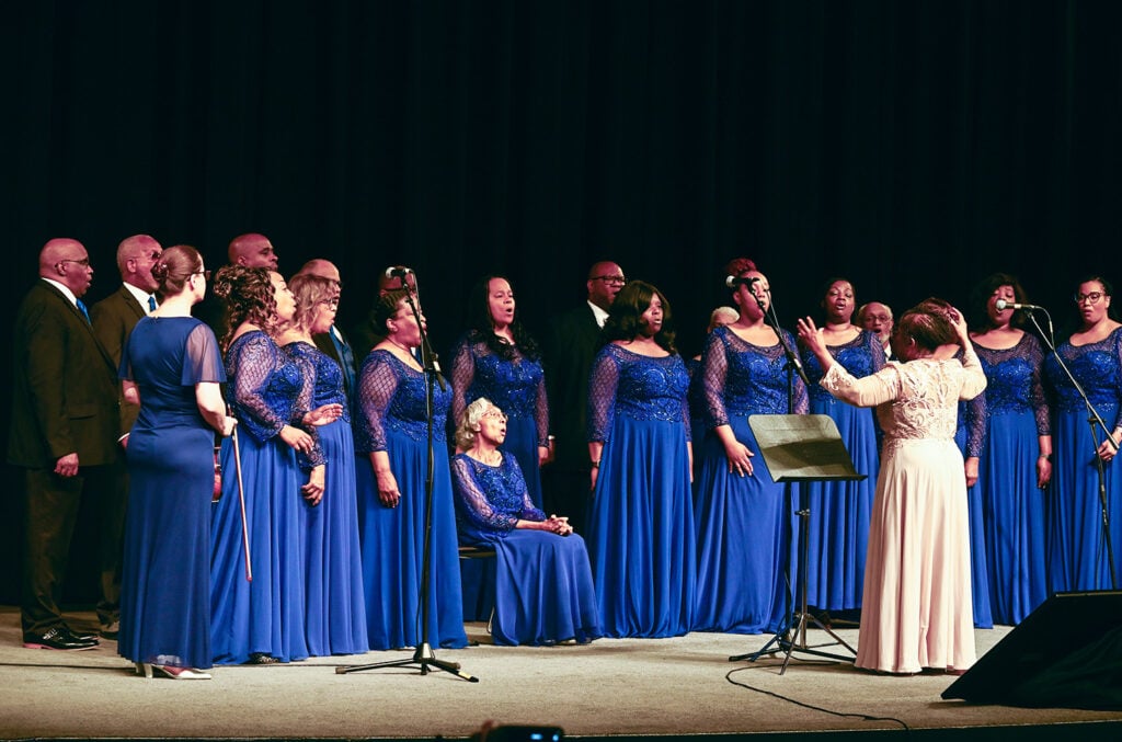 A choir of male and female singers performs under a female conductor. One choir member is seated on a chair. 