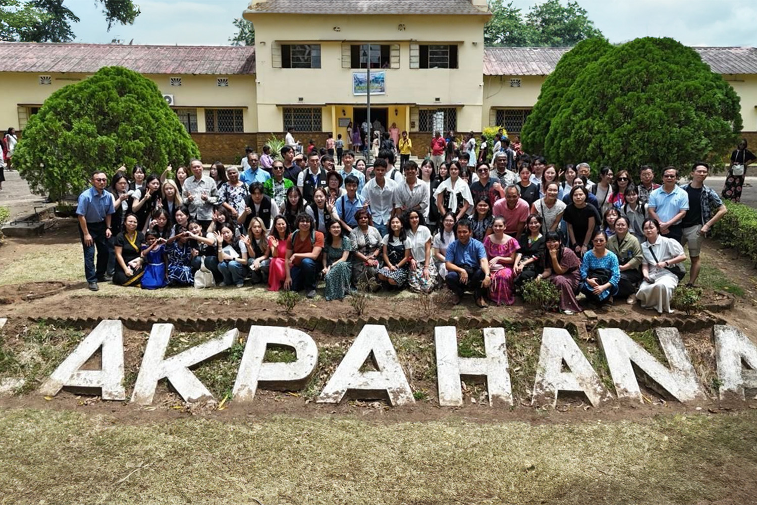 Large group photo of students, faculty, and visitors gathered in front of a college campus. The group poses together in rows, with 'LAKPAHANA' spelled out in large white letters on the ground in front of them.